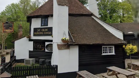 A white building with black painted around the windows. There are two signs with "Ye olde fighting cocks" on them. Trees are in the background.