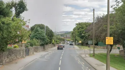 A road with walls and houses on the left hand side. On the right, trees and lampposts. A bridge in the distance.