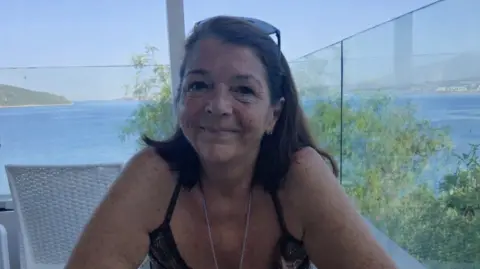 Julie Wilson is pictured with shoulder-length brown hair sitting at a table on a terrace overlooking the sea, smiling at the camera, with blue water and a distant coastline visible behind her.