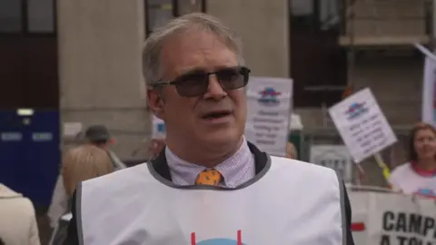 Protester Scott Slavin wears sunglasses and a white tabard in front of a group of protesters carrying signs and placards.