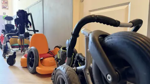 Different mobility aids and wheelchairs in the corridor of Izzy's family home.
