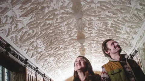 Steve Haywood Two people looking up at the ceiling which has different moulds with different depictions.