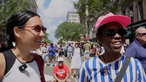 Two women wearing sunglasses stand talking in a street with microphones clipped to their shirts.