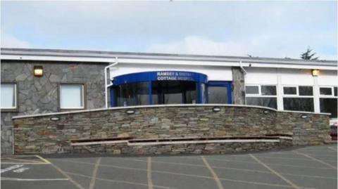 Ramsey Cottage Hospital's main entrance. A stone-built wall sits in front of the entrance which is a round revolving door with a blue frame and the words Ramsey & District Cottage Hospital written in white lettering above the door. The building itself is stone-clad on the left hand side, and white on the right hand side, with both sides featuring windows. A car park with yellow criss-cross lines on it can be seen in the foreground.