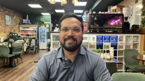 Man with glasses and a beard looking into the camera, sitting at a table inside a cafe