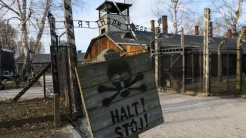 An exterior view of the Auschwitz concentration camp, with a wooden sign in the foreground with the word "halt" on it. Barbed wire fences sit behind the sign