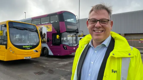 Doug Claringbold wearing black rimmed glasses, a blue shirt and a black blazer underneath a hi-vis yellow jacket. He is smiling at the camera, and standing in the Weston bus depot in front of two diesel buses which will soon be replaced with electric ones. 