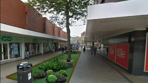 The low brick buildings, with their white overhanging covers face each other down a long narrow shopping precinct. A computer generated image has been added to the centre of the pedestrianised walkway showing a tree and plant beds.