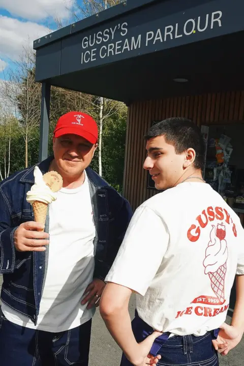 Gussy's Ice Cream Two people stand outside Gussy’s Ice Cream Parlour. Gus holds a soft-serve ice cream cone, while his son Oli wears a white T-shirt featuring a large red ice cream graphic and the words “Gussy’s Ice Cream Est. 1959” on the back.