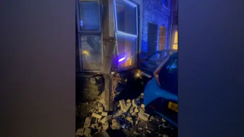 Msadiq Hussain Damage to the front bay window of a brick house shows masonry falling off and a pile of bricks on the pavement. Two cars can also be seen.