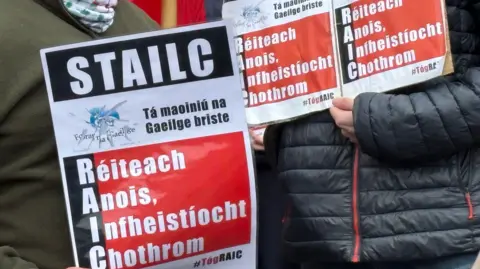 PA Media Workers from Irish language groups hold signs as they protest outside Culturlann McAdam O Fiatich, in west Belfast as their colleagues across Ireland take part in strike action over funding cuts in the sector.