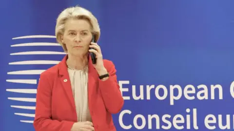 A woman in a pink jacket in front of a blue background holds a mobile phone to her ear