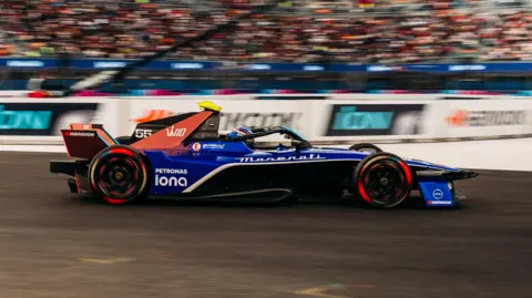 Maserati MSG Racing A blue and red racing car on a racing track with the audience and advertising boards blurred in the background.