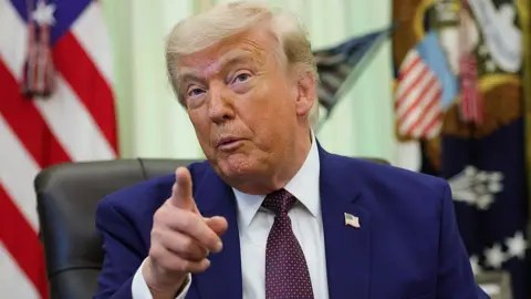 Donald Trump sits in a leather chair in the Oval Office and points his index finger while speaking