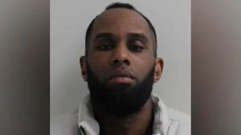A mugshot of a man with a wearing a white coat with a dark beard and short hair.