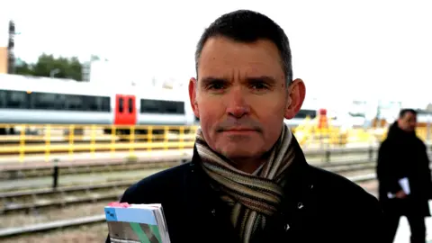 Qays Najm/BBC David Hughes, wearing a dark coat and a colourful, striped scarf tied neatly round his neck, looking down the camera. He is standing outside, next to some railway lines.