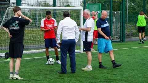Surrey Heartlands NHS A group of male football players have a conversation in front of a white goal. They are chatting on a green football pitch made of artificial grass.