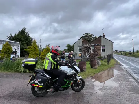 A motorbike being ridden by a man wearing a high vis jacket and red helmet past a cafe called "Silver Fox" There is a white double decker bus in the background parked near a lawn with some trees. 