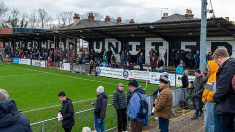 Supporters at Maidenhead United stadium, with a stand painted black and white with Maidenhead United painted on it 