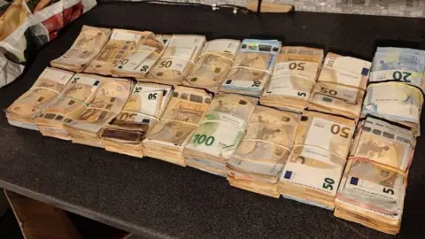 An Garda Síochána مجموعات من الأوراق النقدية من فئة 50 و100 يورو موضوعة في صف واحد على سطح طاولة داكن اللون