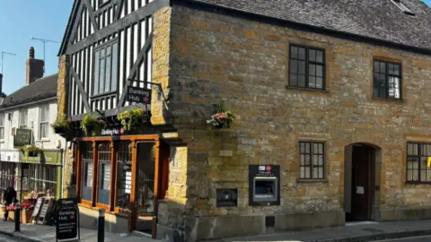 Large stone building with cash machine in its side wall, a sign on the pavement outside and it's door open.  The are several hanging baskets along its walls.