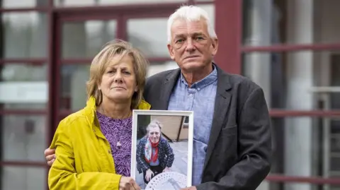 PA Media A man and a woman standing beside each other. They are holding a framed photo of a woman. 