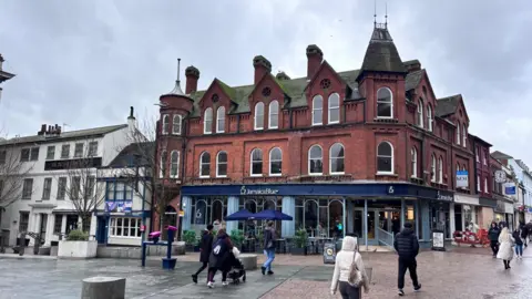 A three-storey red brick building with chimneys and turrets is at the side of an open, pedestrianised square in a town centre. A pedestrianised street runs along the side of the building on the right. There is a pub and hotel building next to it on the left. Shoppers are walking in front of it.