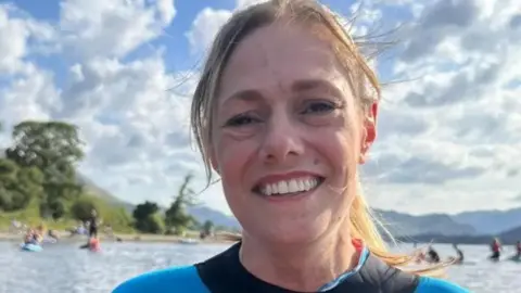 Photograph of cancer campaigner Kate Rackham - she is in a wet suit with water behind her and people canoeing behind her. Hills and trees are also behind her surrounding the water. 