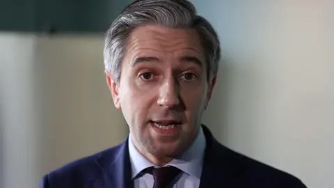 Simon Harris is wearing a blue shirt, navy tie and navy blazer. He has grey hair. The background is blurry. 