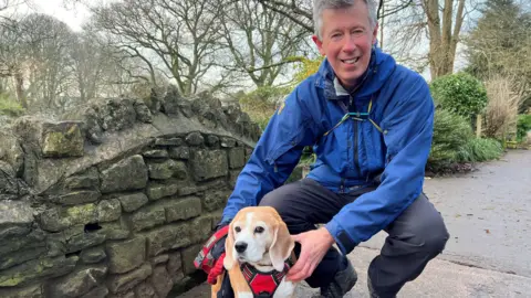 A man in a blue coat leaning down to his pet dog. It's a small beagle on a bridge in a park with lots of trees in the background.