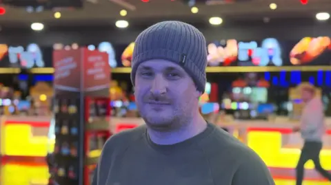 Connor Langley wearing a grey-coloured beanie hat, standing in front of a yellow-lit food counter inside the cinema.