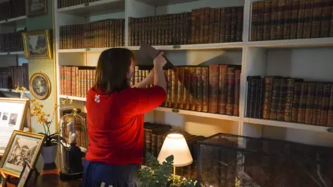 A National Trust staff member examines a row of old volumes on wooden shelves at Stourhead with other curiosities around her and small lamp. We see her back and she wears a red fleece with the trust logo.