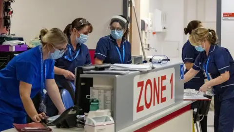 stock image of Whiston Hospital workers in their blue uniforms and surgical masks at a reception desk with a sign saying Zone 1