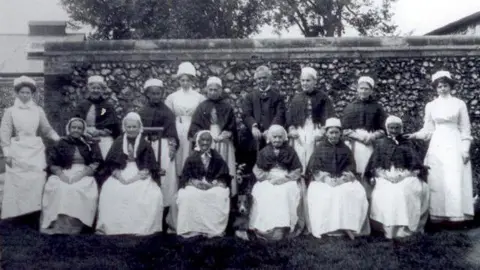 Thetford Town Council Dr Allan Minns, wearing a dark three piece suit, standing with 11 Thetford Union Workhouse women inmates and three nurses. He is in the back row, standing, wearing a dark three piece suit. The women inmates are wearing shawls over light-coloured dresses and light coloured caps on their hair. There are also three nurses, two at either end and one standing towards the middle