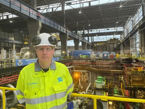 Peter O'Grady wears a yellow hi-visibility jacket, safety googles and a white hard hat. He is stood in front of yellow railings in a building at the power station.