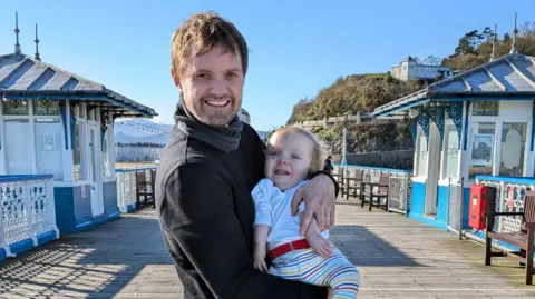 Harvie Strange Barney Strange holding Rafferty in his arms. He is standing on a promenade and they are both smiling for the camera.