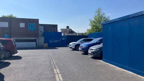 Large wooden walls painted blue installed in a car park. There are cars parked in spaces, and a sign for Co-op in the distance.