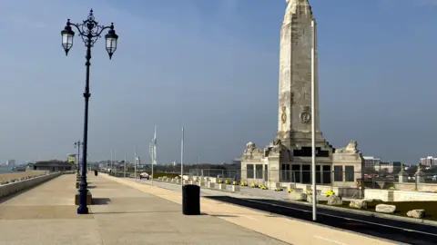 Portsmouth City Council Portsmouth Naval Memorial is a tall, white stone monument with four sides. It is beside a wide, paved promenade with ornate lamp-posts.