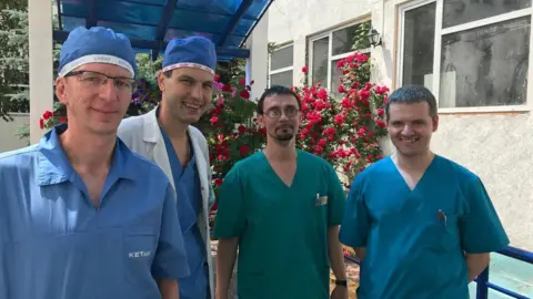 Leo's Row Four men stands outside a hospital wearing scrubs. Leo is on the left, wearing light blue scrubs and a matching cap and thin wire framed glasses. To the right, a man with black hair wears pale blue scrubs, a blue cap and a white overcoat, a man stands next to him with black short hair and a black beard, wearing deep green scrubs and another man stands next to him, with matching blue scrubs on. They are all smiling at the camera