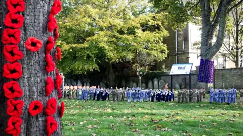 Mandy Wright Photography Tree bark covered with red knitted poppies with knitted small soldiers placed on the grass in the distance