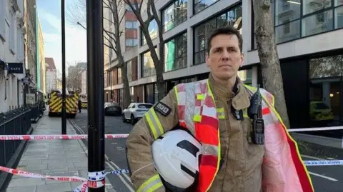 Alec McNally, station commander at the LFB, at the scene. He is wearing his firefighters uniform and carries a helmet.