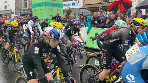 A group of cyclists in racing gear ride closely together on a wet town street during a cycling event. Several riders wear bright helmets and team-branded jerseys, while spectators stand behind barriers holding umbrellas and watching from the roadside. The road surface appears rain-soaked, and sponsor signs and shopfronts are visible in the background