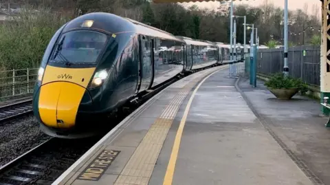 A green GWR train pulls into a railway station. It has its lights on. The platform is empty.