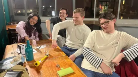 Four students sitting around a wooden table. They are laughing and posing with one elbow up, pressing their fingers down on the table as if they are pressing a buzzer