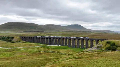 Avanti West Coast/PA A train running along the Settle to Carlisle railway line over the Ribblehead Viaduct. The stone viaduct with its many arches is surrounded by rolling green land with fells in the background.
