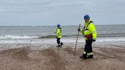 HM Coastguard Two HM Coastguard employees walking on a beach, both wearing high vis and blue helmets, looking for nurdles - small plastic pellets. The one closest to the camera is holding a long stick.