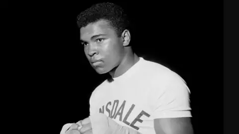 PA Media Black and white portrait of a boxer, Muhammed Ali, in a fitted T-shirt with “Lonsdale” printed across the chest, hands wrapped and raised in a guard position, standing against a dark background.

