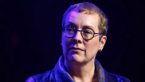 Getty Images A close-up of Julia Angwin. She is wearing round, dark-rimmed glasses and a dark leather jacket, looking slightly off-camera. She has short, light-brown hair, and the background is dark with soft blue and purple lighting.