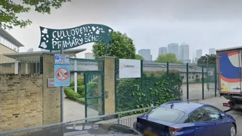 Google Image showing the entrance to Culloden Primary School, with light-coloured brick walls, grey railings and green metal fencing, and a green wavy sign with the school name cut out of it