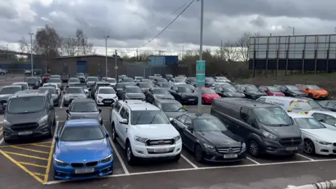 A crowded outdoor car park with various cars and vans under an overcast sky